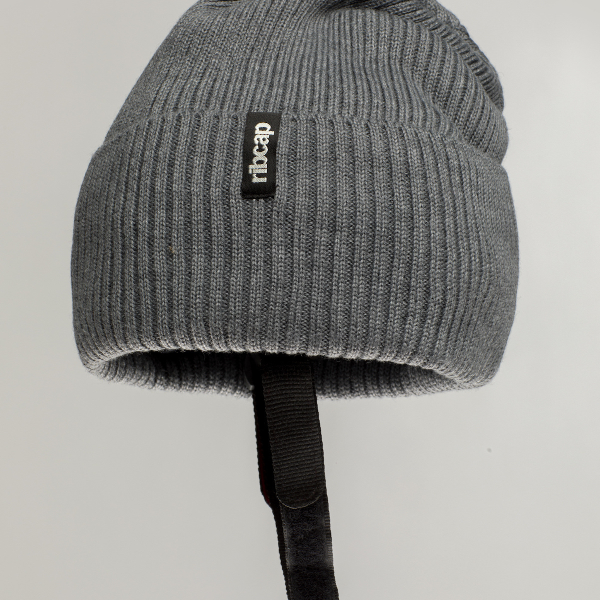 The Ribcap Lenny Beanie Protective Helmet [Grey] by Ribcap is a gray knit helmet for adults, featuring a black rectangular "cheap" tag and a black strap underneath, showcased on a plain light background.