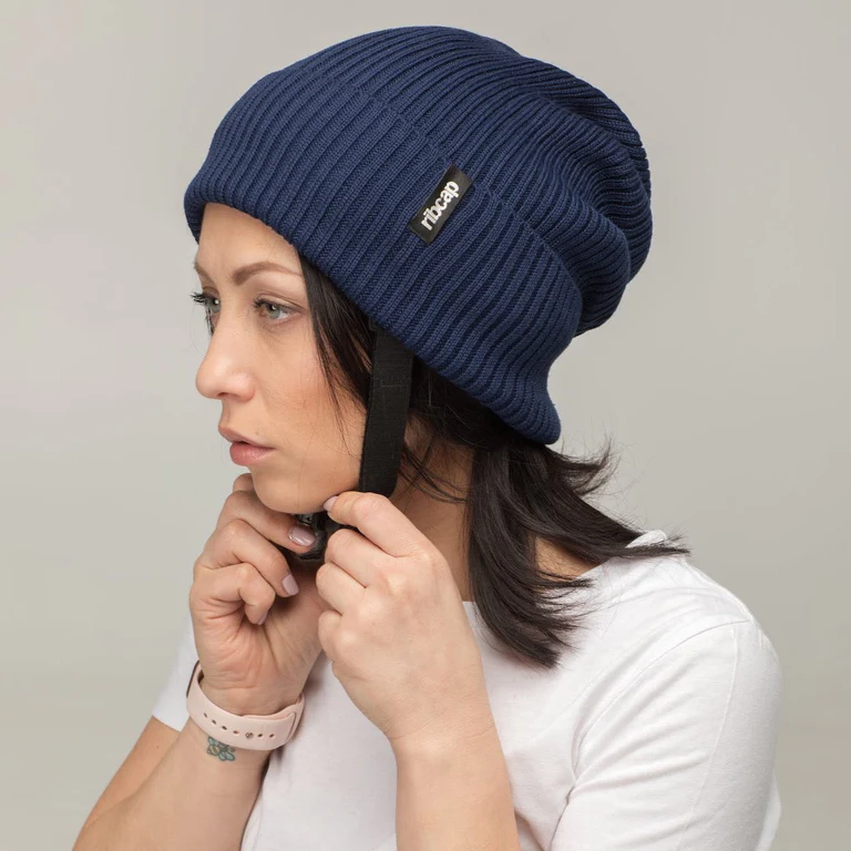 A woman in a white shirt adjusts the black chin strap of a Ribcap Lenny Beanie Protective Helmet [Marine] by Ribcap. The blue adult knit beanie features a black label, and she gazes slightly left against a plain background.