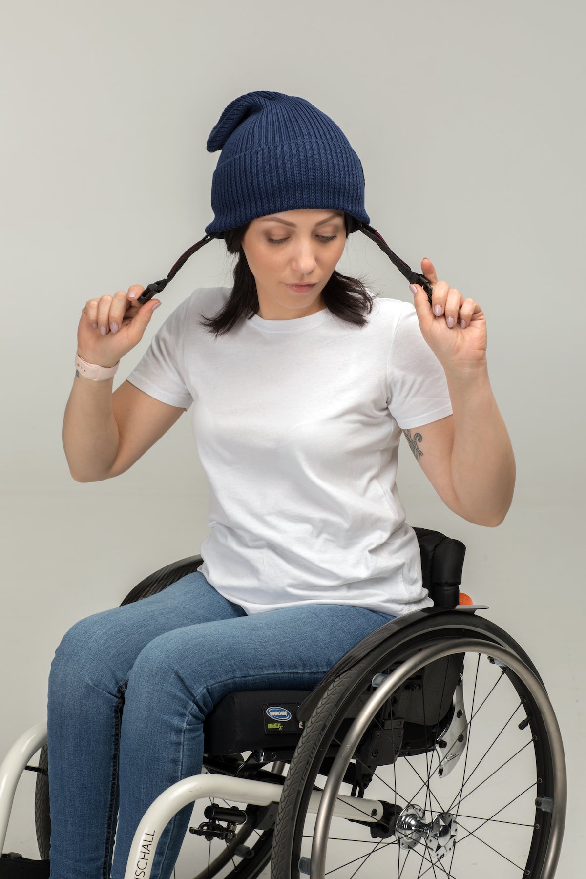 A woman in a wheelchair wears the Ribcap Lenny Beanie Protective Helmet [Marine] by Ribcap, along with a white t-shirt and jeans, holding her braided hair ends against a plain light background.