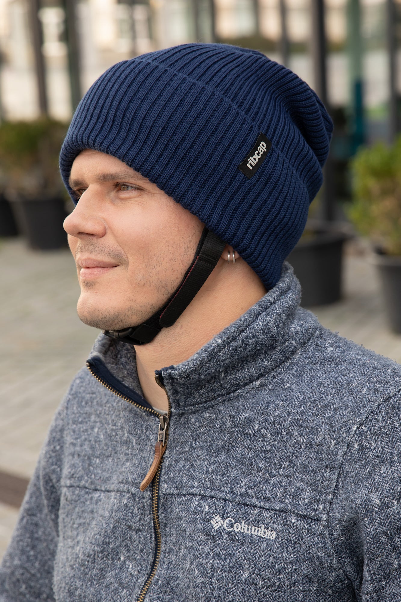 A man wears a Ribcap Lenny Beanie Protective Helmet [Marine] by Ribcap with a black strap and a gray Columbia zip-up jacket, standing outdoors in front of potted plants and a blurred background.