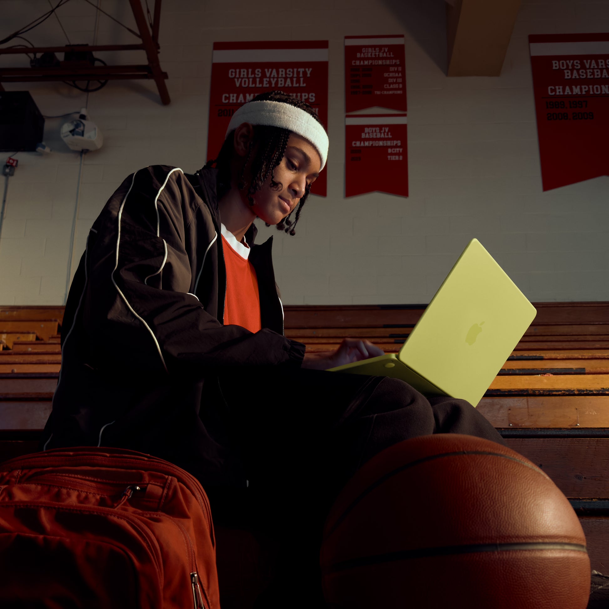 A person in a beanie and tracksuit sits on gym bleachers, typing on an Apple MacBook Neo with Liquid Retina display. A backpack and basketball are nearby, and sports banners hang on the wall.