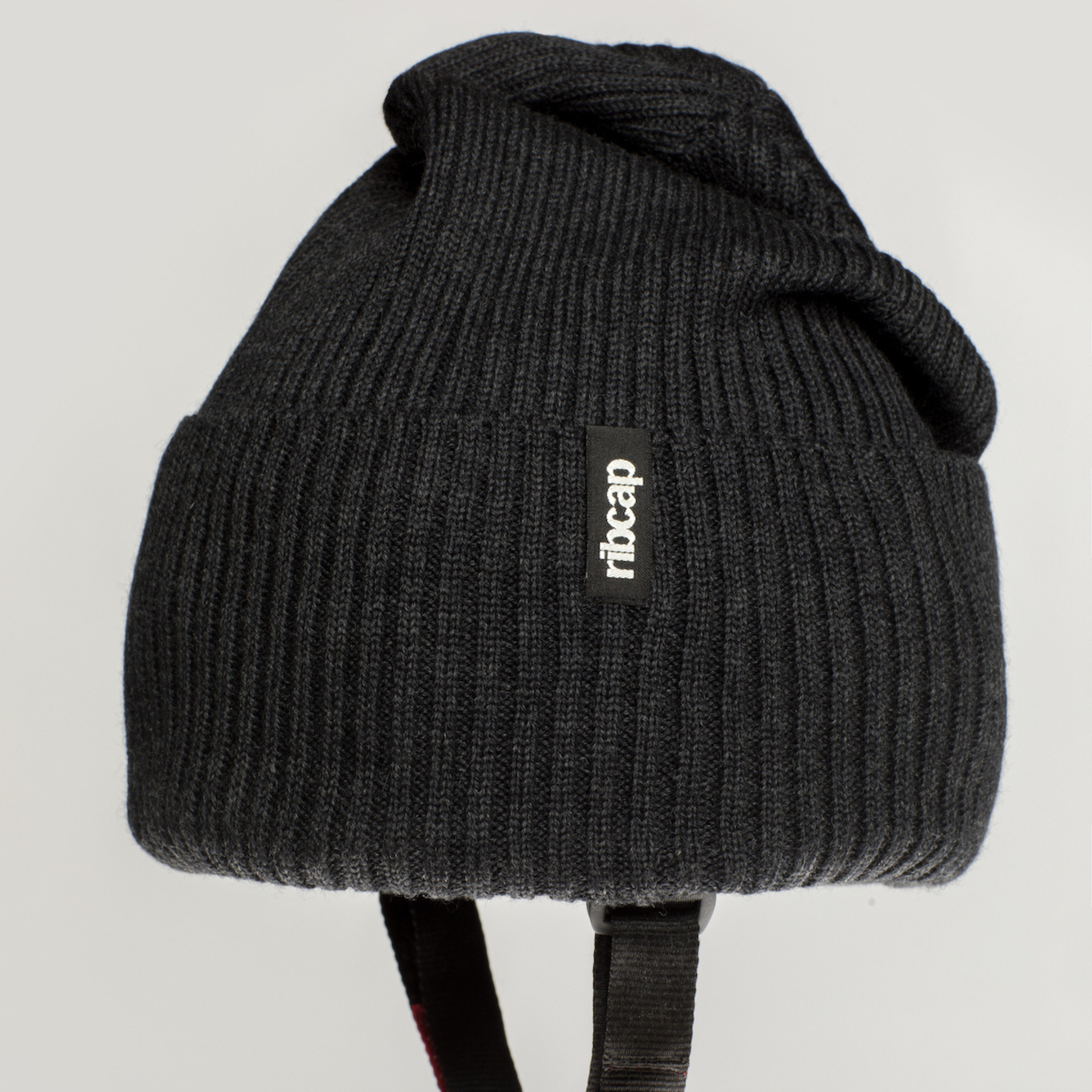 The Ribcap Lenny Beanie Protective Helmet [Anthracite] by Ribcap features black ribbed knit, a small front label, and black chin straps. Shown against a plain light grey background.