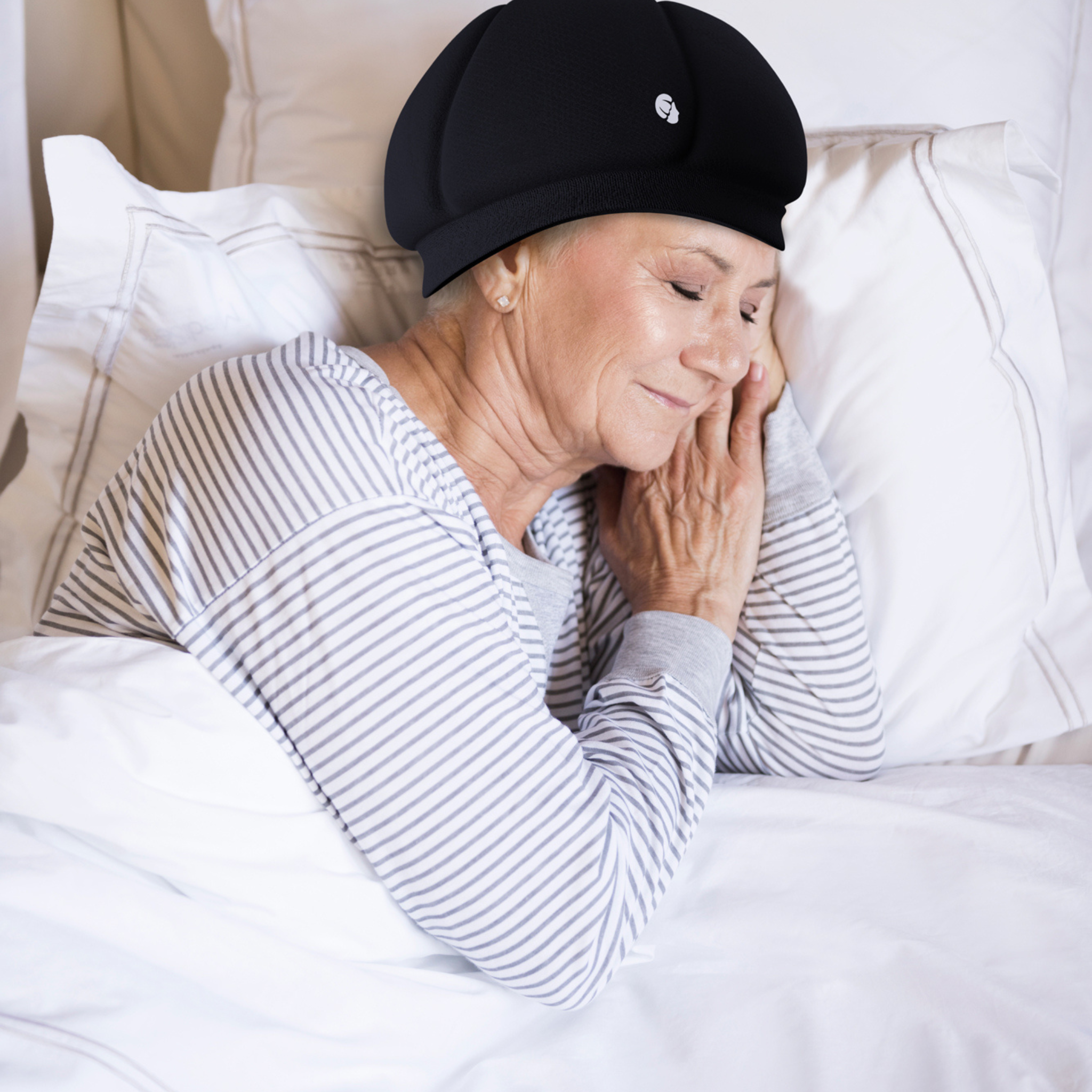 An older woman in a striped pajama top and the Ribcap Kate SleepSafe Cap [Black] by Ribcap rests peacefully on her side in a neat white bed, smiling gently with her head supported by a pillow.
