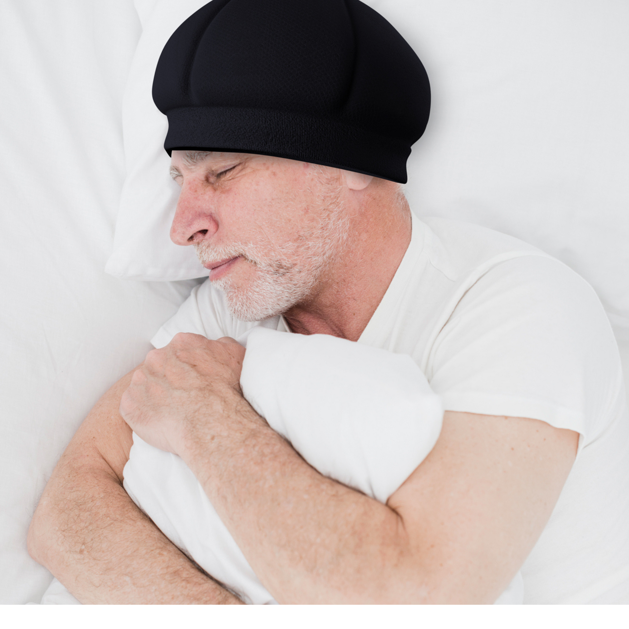 An older man with a white beard sleeps on his side, hugging a white pillow and wearing the Ribcap Kate SleepSafe Cap [Black] by Ribcap, dressed in a white t-shirt on white bedding.