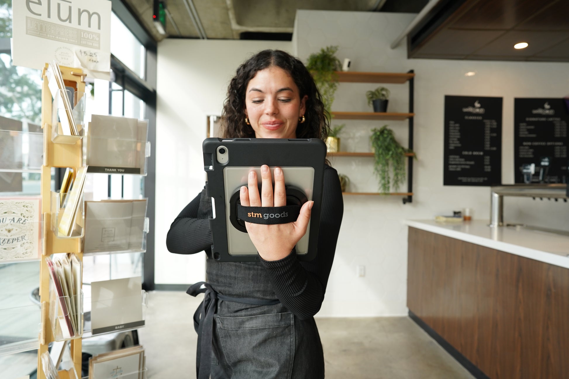 A woman in a modern café holds an STM Swivel Case [Black] (for iPad A16/10th/11th Gen) by STM, smiling as she grips her tablet with the strap. Plants, shelves, and a menu board are visible in the background.