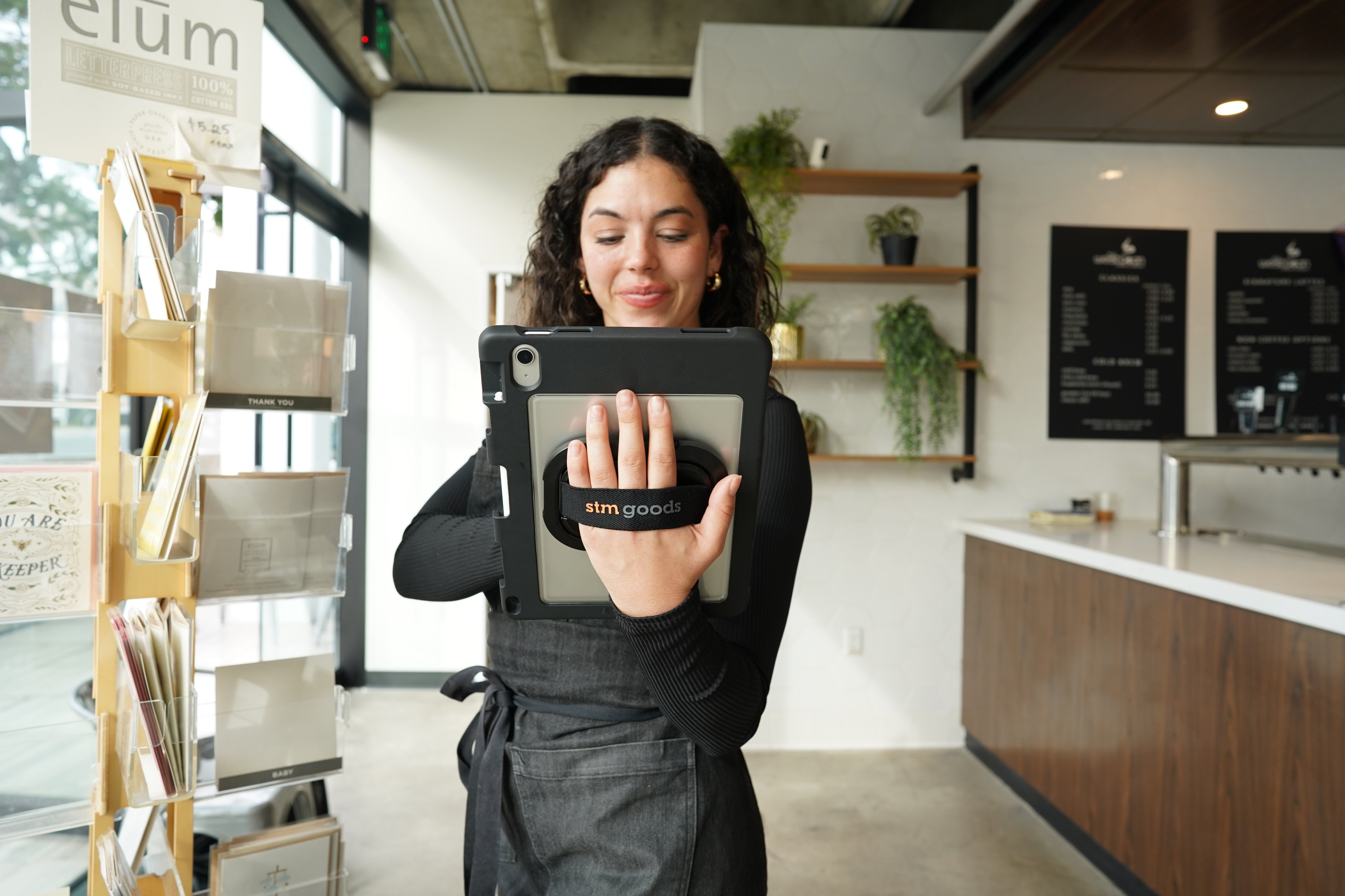 A woman in a modern café holds an STM Swivel Case [Black] (for iPad A16/10th/11th Gen) by STM, smiling as she grips her tablet with the strap. Plants, shelves, and a menu board are visible in the background.