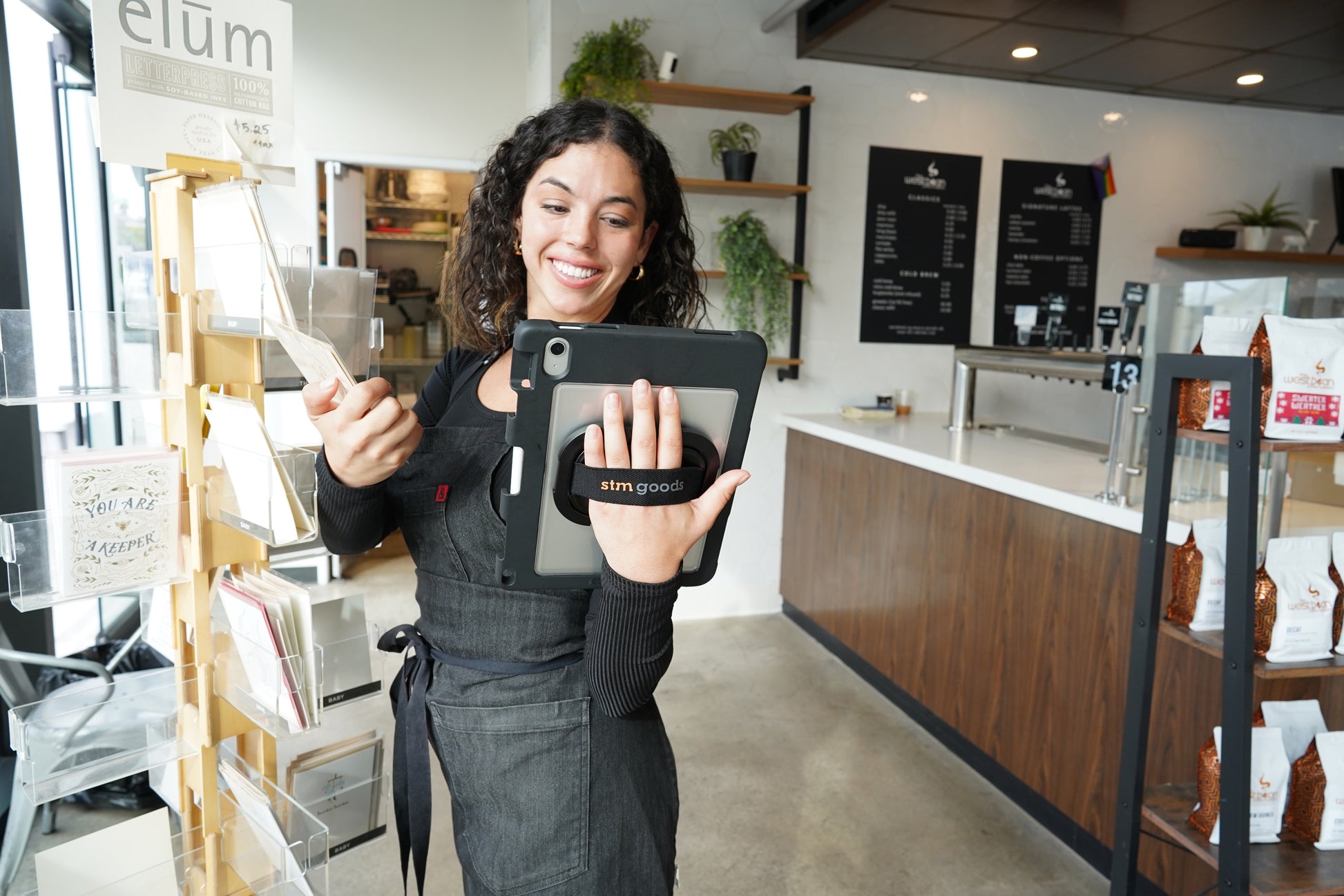 A smiling woman in an apron holds an STM Swivel Case [Black] for iPad by STM and reaches for a greeting card in a bright modern café, with a visible menu board, coffee bags, and plants in the background.