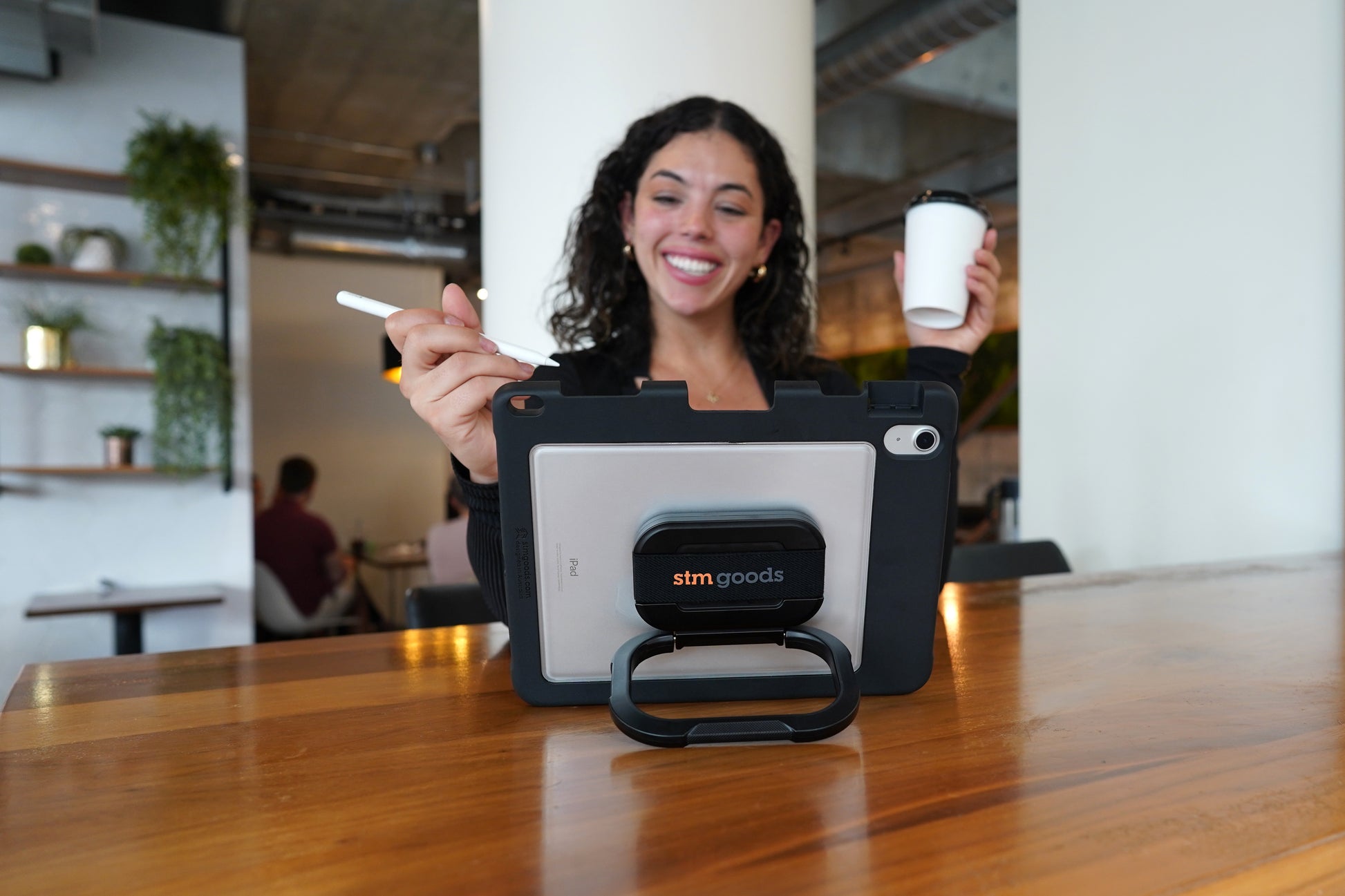 A woman smiles as she uses her iPad in an STM Swivel Case [Black] (for iPad A16/10th/11th Gen) by STM, holding a stylus and coffee at a wooden table indoors with shelves and plants in the background.