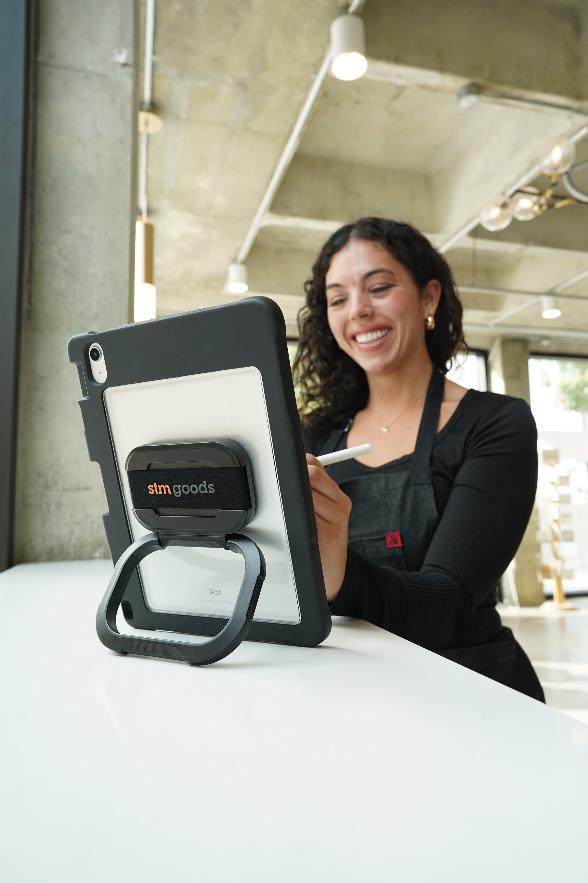 A smiling woman in a black shirt and apron stands at a counter using an iPad in the STM Swivel Case [Black] (for iPad A16/10th/11th Gen) by STM, in a modern, well-lit space.