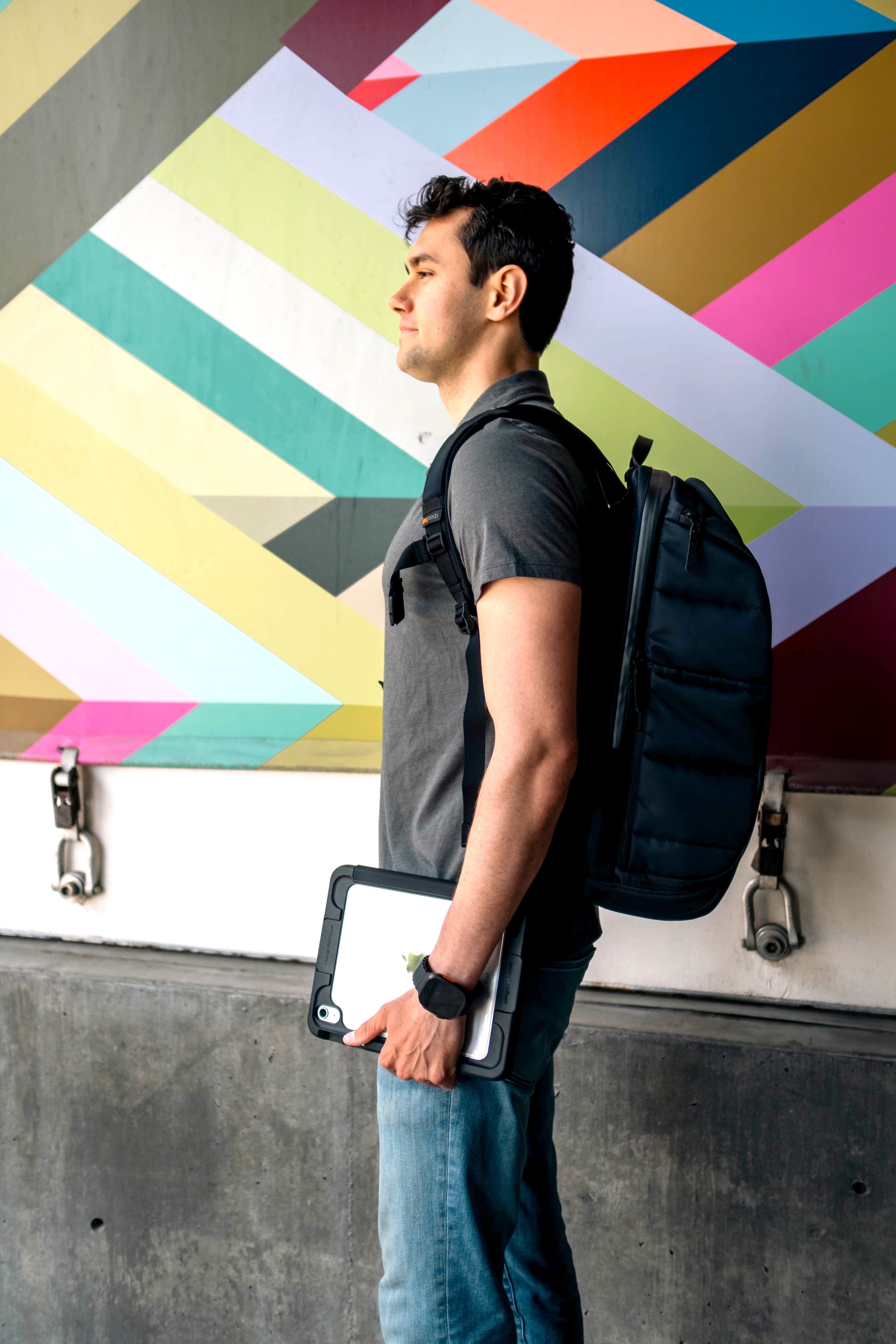 Man with a backpack holding a tablet in front of a colorful geometric wall.