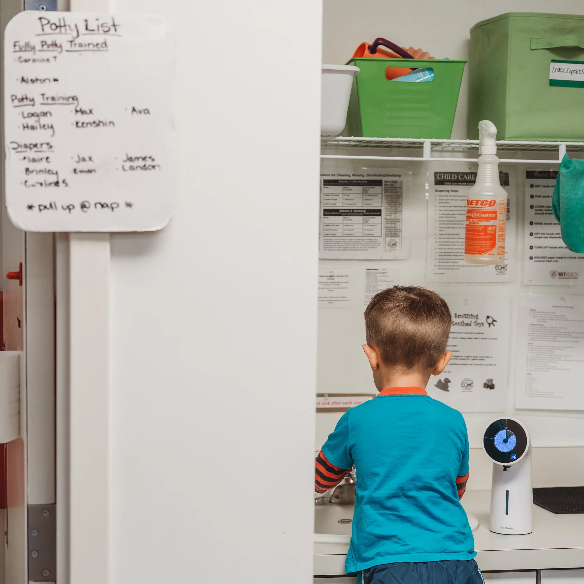 A young boy washes his hands in a classroom using the Time Timer PRO Applied Soap Dispenser Timer by Time Timer, with a Potty List on the whiteboard and shelves with supplies in the background.