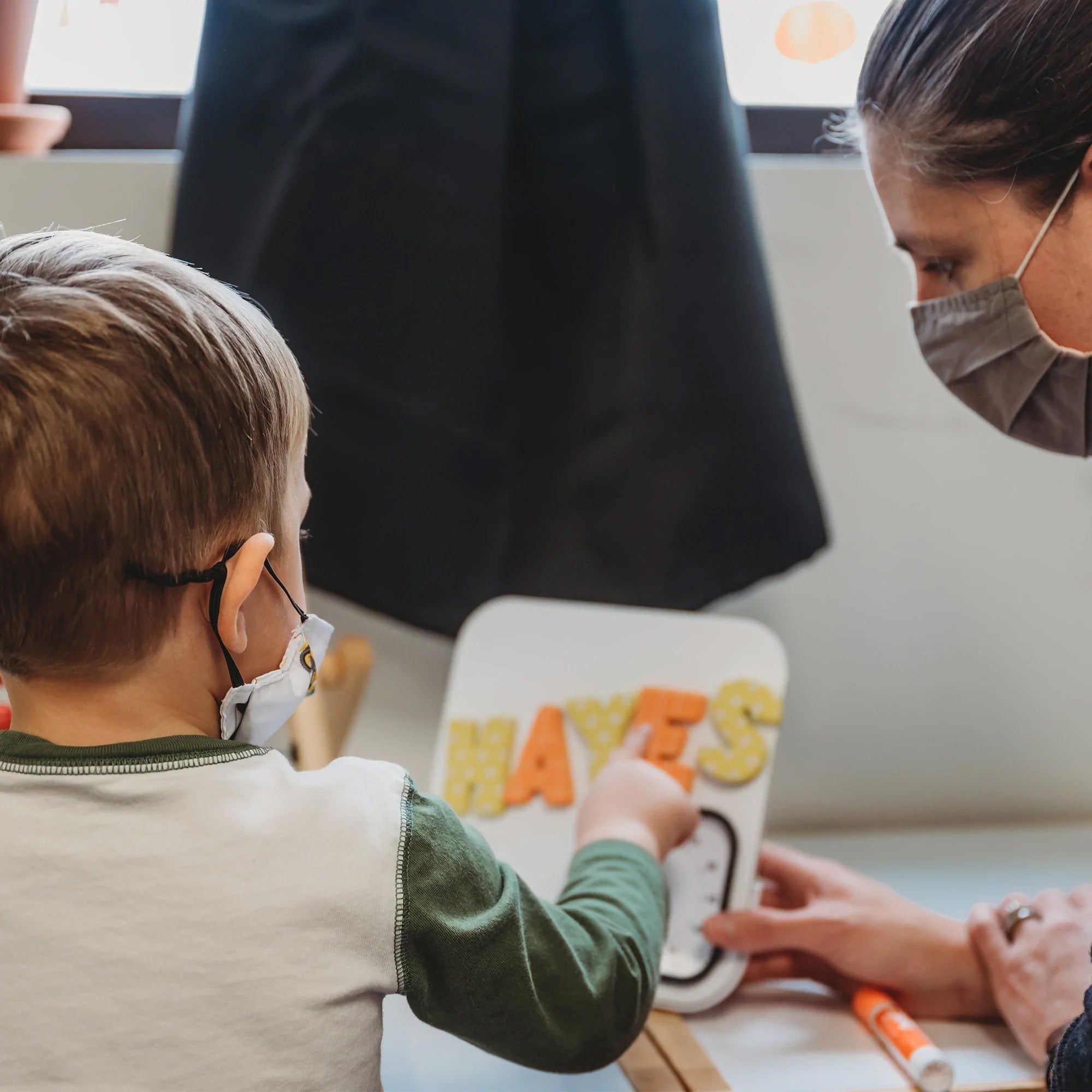 A young child and an adult, both masked, sit at a table. The child points to the Time Timer MOD with Magnetic Whiteboard (60 min) by Time Timer, where "HAYES" is written in colorful letters as the adult observes and assists.