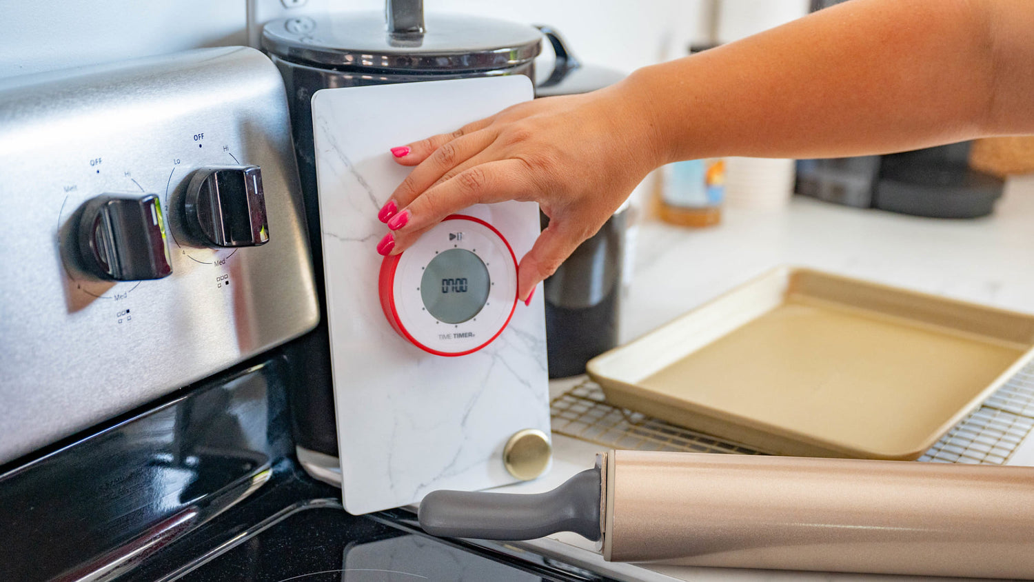 A hand with pink nail polish sets a digital kitchen timer on a marble-patterned stand next to a stovetop, with a baking sheet and rolling pin nearby on a countertop.