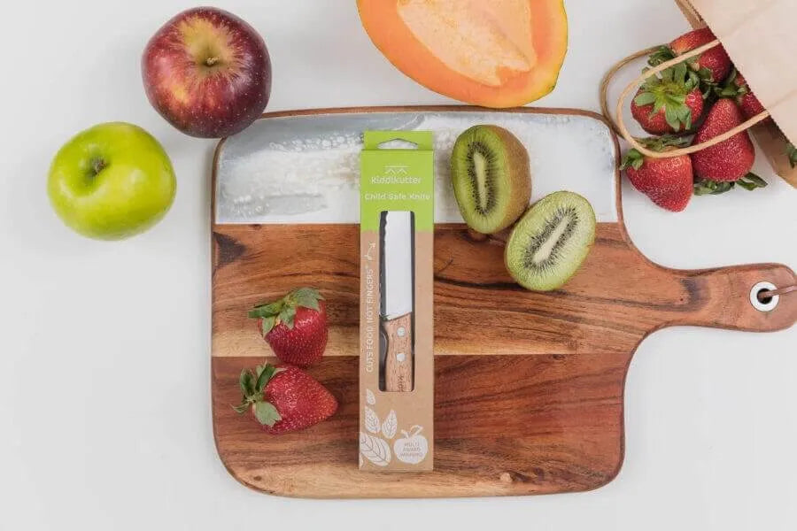 A wooden cutting board displays apples, strawberries, kiwi, and cantaloupe. On it is a KiddiKutter Kid Safe Knife [Wooden] by KiddiKutter. A paper bag filled with strawberries sits close by.