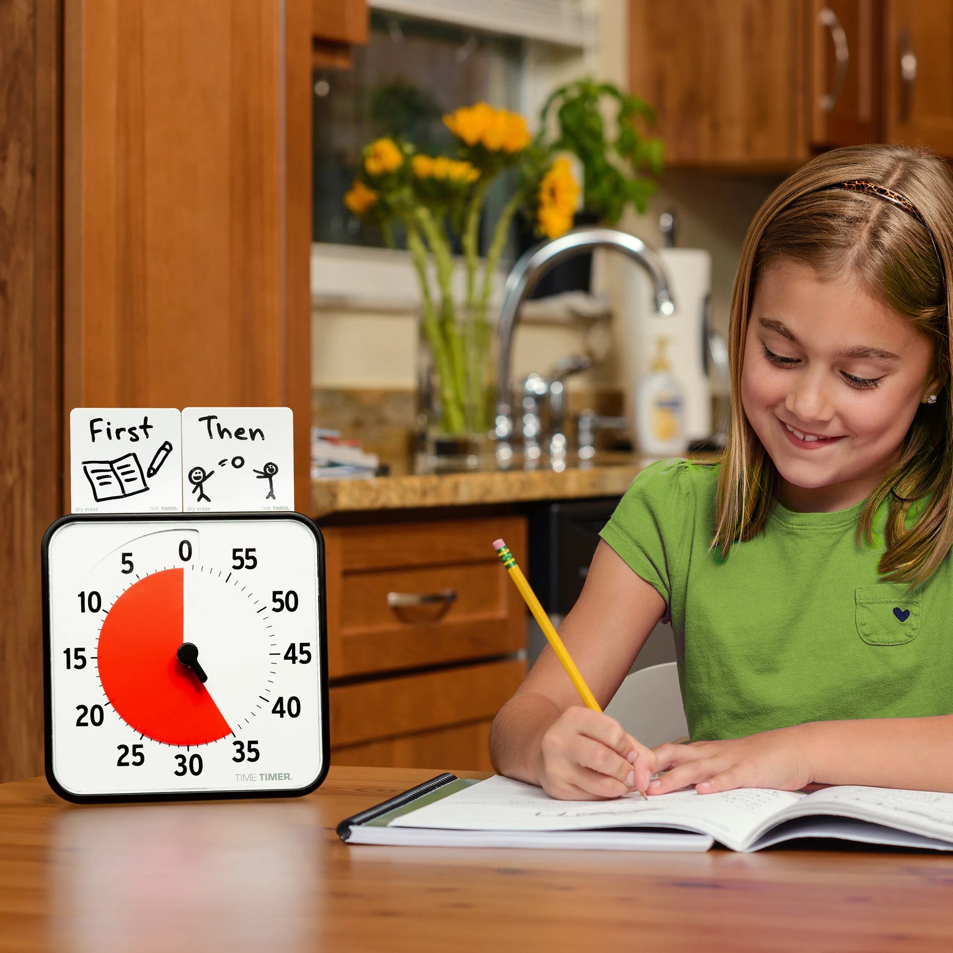 A young girl in a green shirt smiles while writing at the kitchen table. Beside her are a Time Timer Original (8 Inch) by Time Timer and a First-Then chart showing "book" first, then "play outside." Flowers sit on the counter behind her.