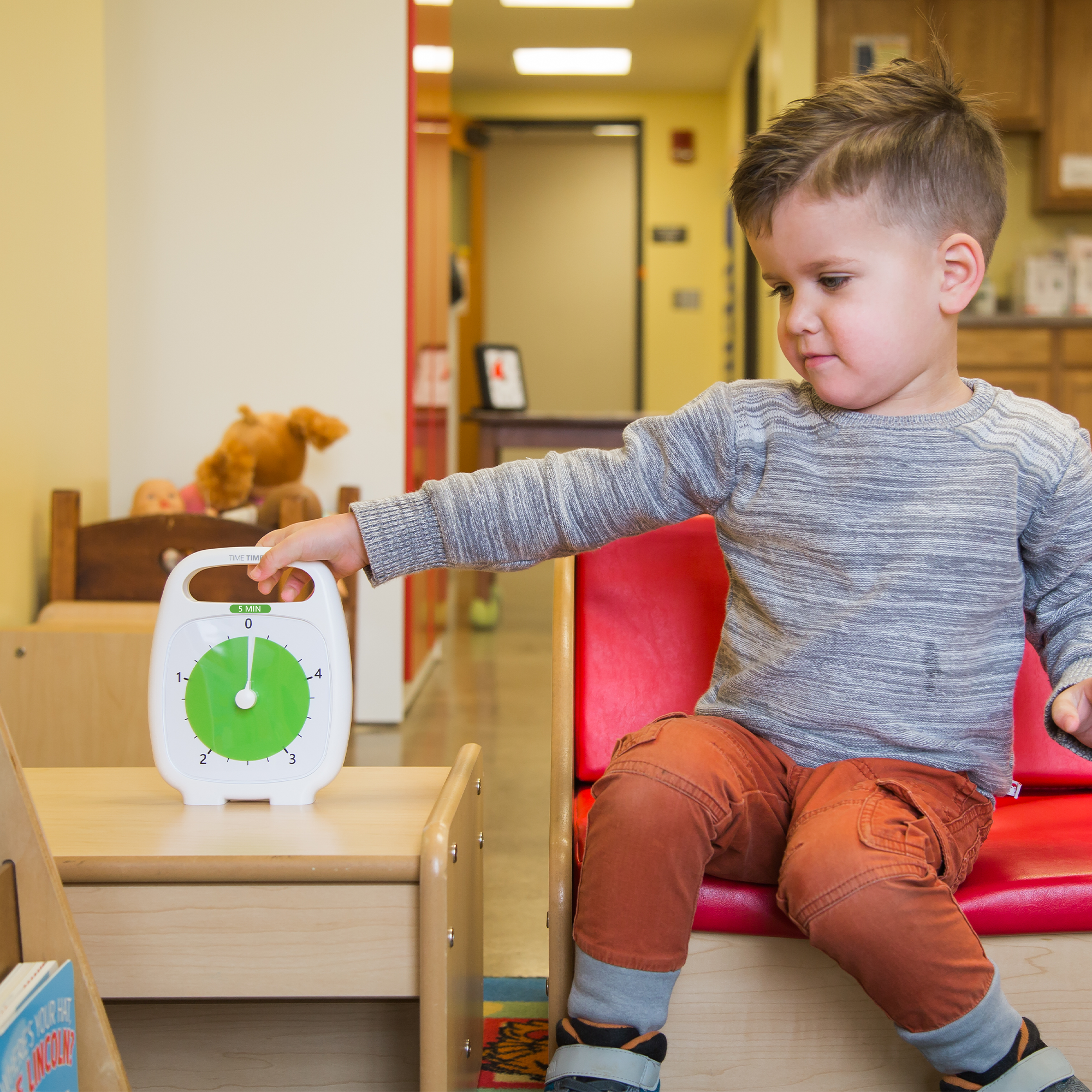 A young boy in a classroom reaches for a white and green Time Timer Plus (5 min) by Time Timer on a low table. The room has wooden furniture, toys, and books in the background.