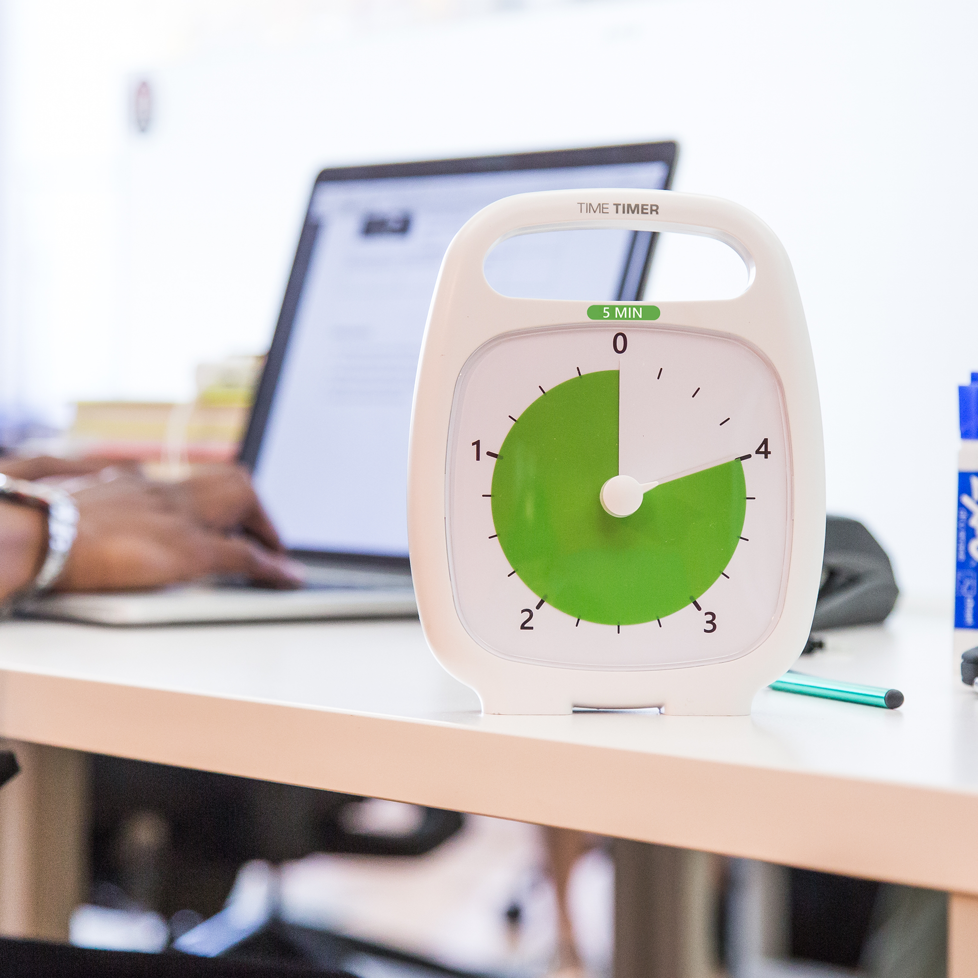 A Time Timer Plus (5 min) by Time Timer sits on a desk beside a laptop while someone types amid office supplies. Its bright, blurred background highlights this visual timer’s focus on keeping you productive and on track.