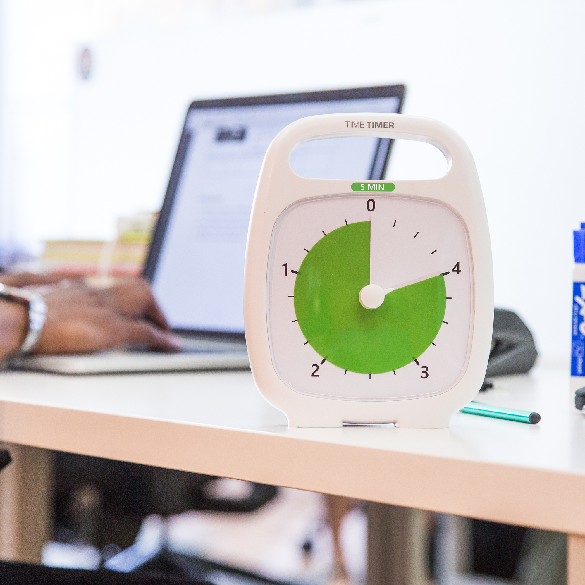 A Time Timer Plus (5 min) by Time Timer sits on a desk beside a laptop while someone types amid office supplies. Its bright, blurred background highlights this visual timer’s focus on keeping you productive and on track.