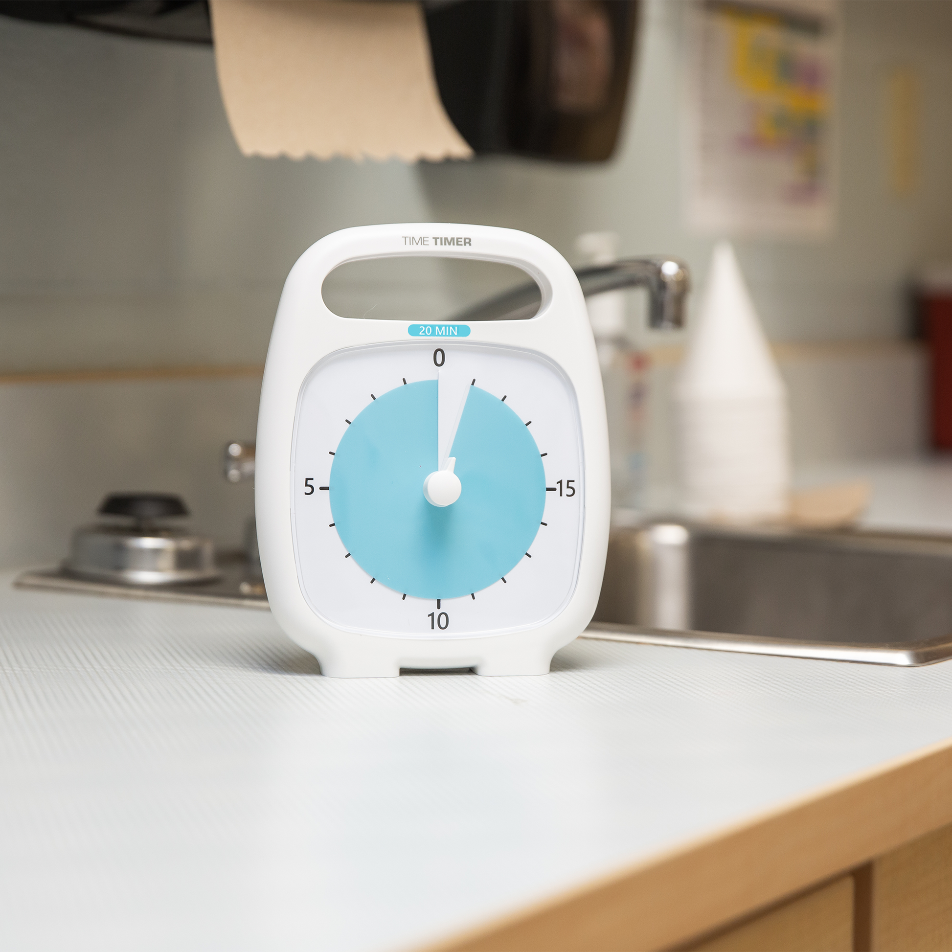 A Time Timer Plus (20 min) by Time Timer, in white and blue, sits on a countertop in a kitchen or break room with a sink and paper towel dispenser in the background, offering an easy-to-see visual timer.