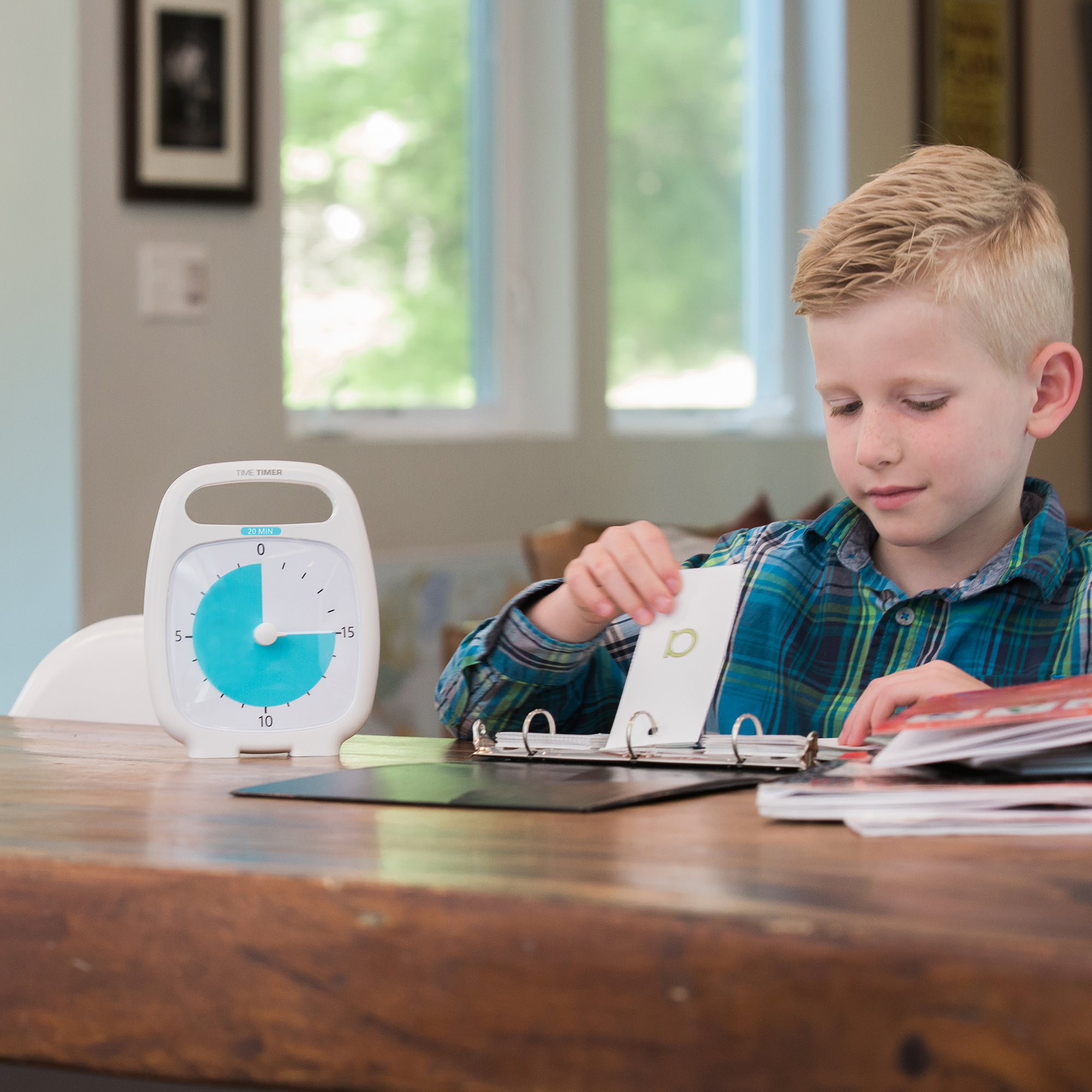 A young boy flips through flashcards at a wooden table with the Time Timer Plus (20 min) by Time Timer beside him. A window and home interior are visible in the background.