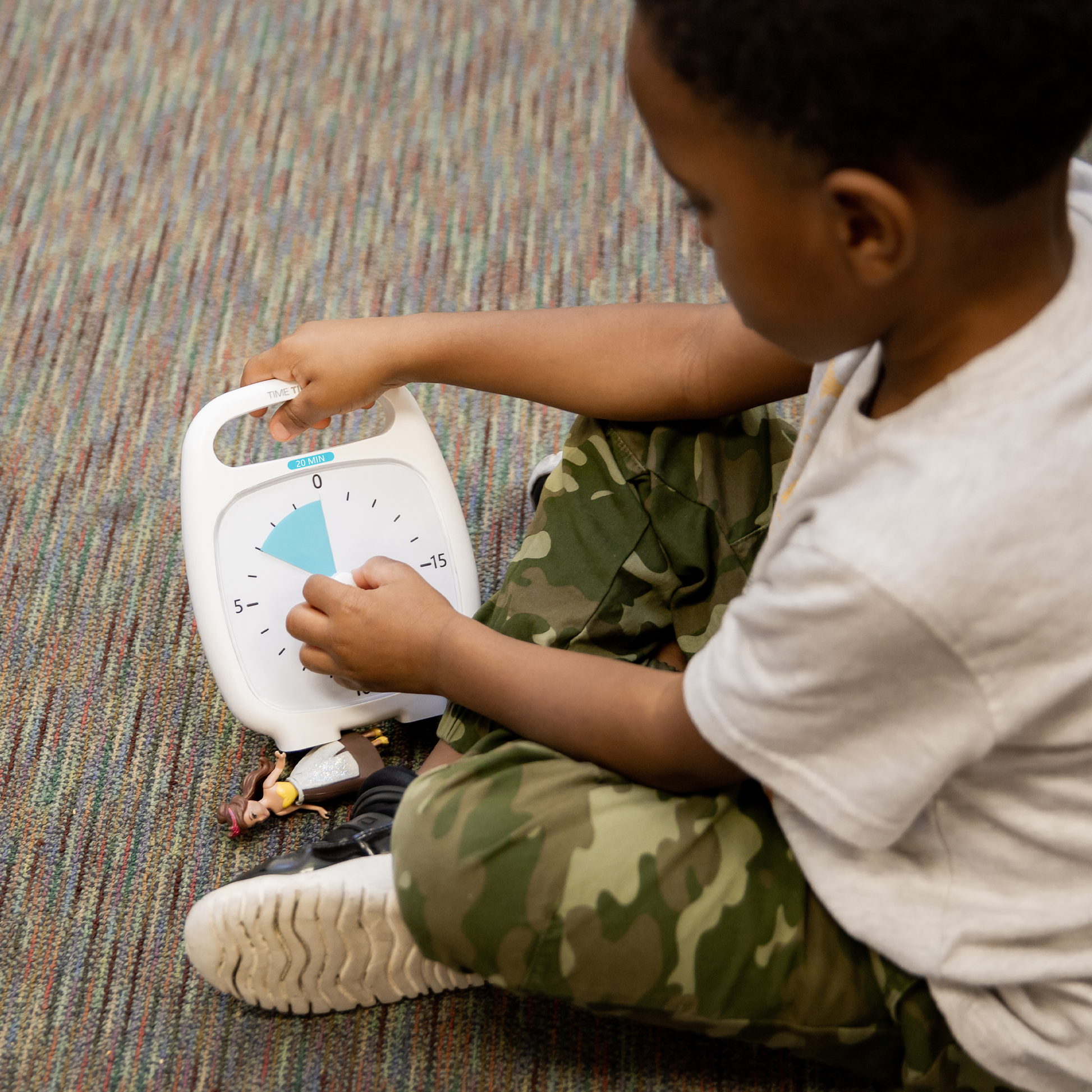 A child in a white shirt and camouflage pants sits on a carpet, setting a Time Timer Plus (20 min) by Time Timer with a blue section. Two small toys are near the child's foot.