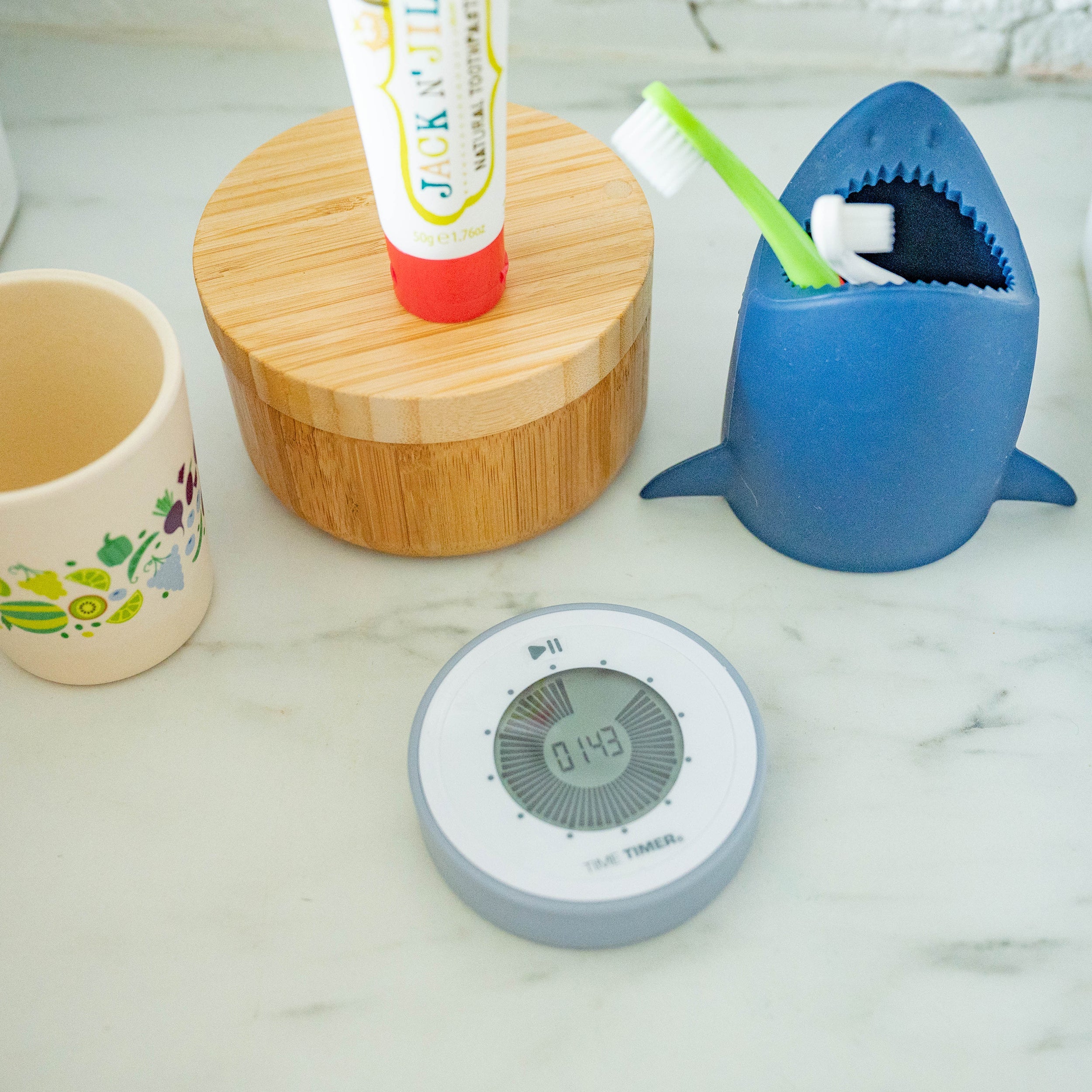 A bathroom countertop featuring the Time Timer Twist by Time Timer (Pale Shale), a colorful cup, a bamboo container with toothpaste, and a shark-shaped toothbrush holder holding a green toothbrush.