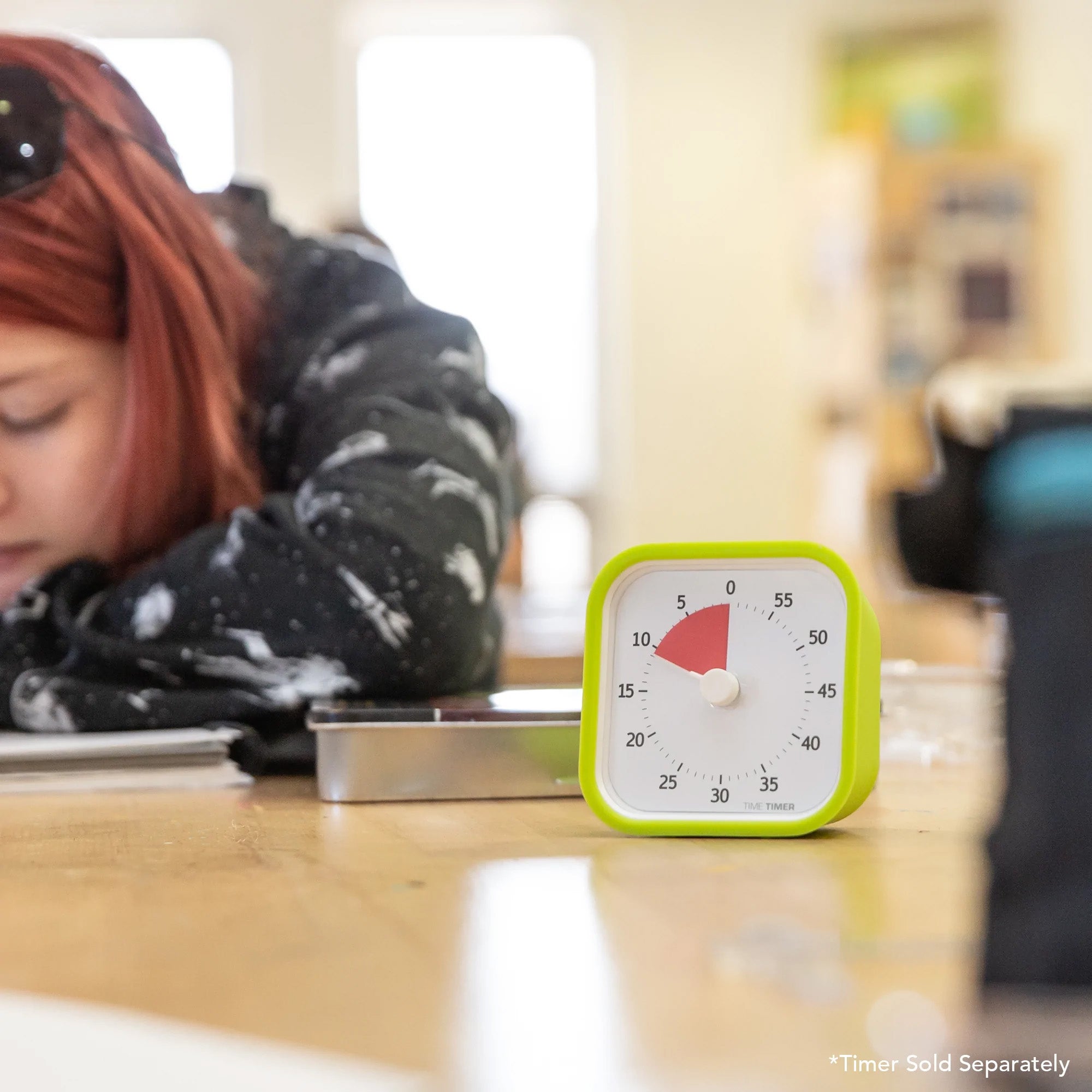 A Time Timer MOD Cover Silicone Case Accessory by Time Timer is in focus on the table, while a red-haired person writes in the blurred background. Caption: Timer Sold Separately.
