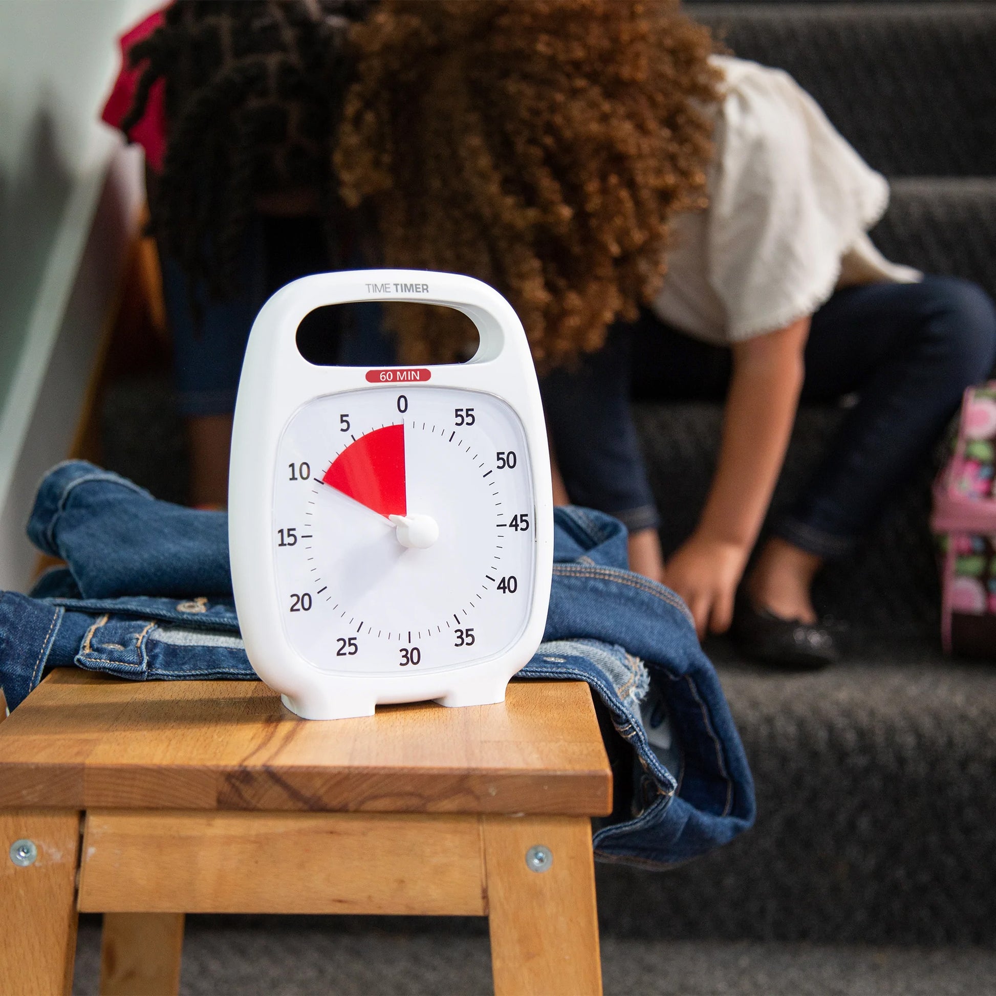 A white Time Timer Plus (60 min) by Time Timer, ideal for time management, rests on a wooden stool draped with jeans. In the background, a child on stairs puts on shoes.
