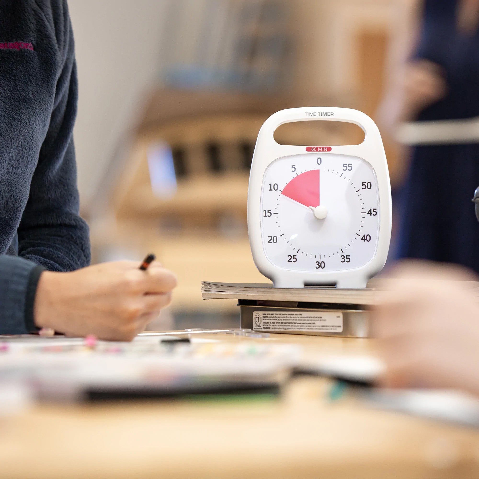 A close-up of the Time Timer Plus (60 min) by Time Timer shows about 19 minutes remaining on a table, surrounded by people writing or drawing. Background objects appear blurred.