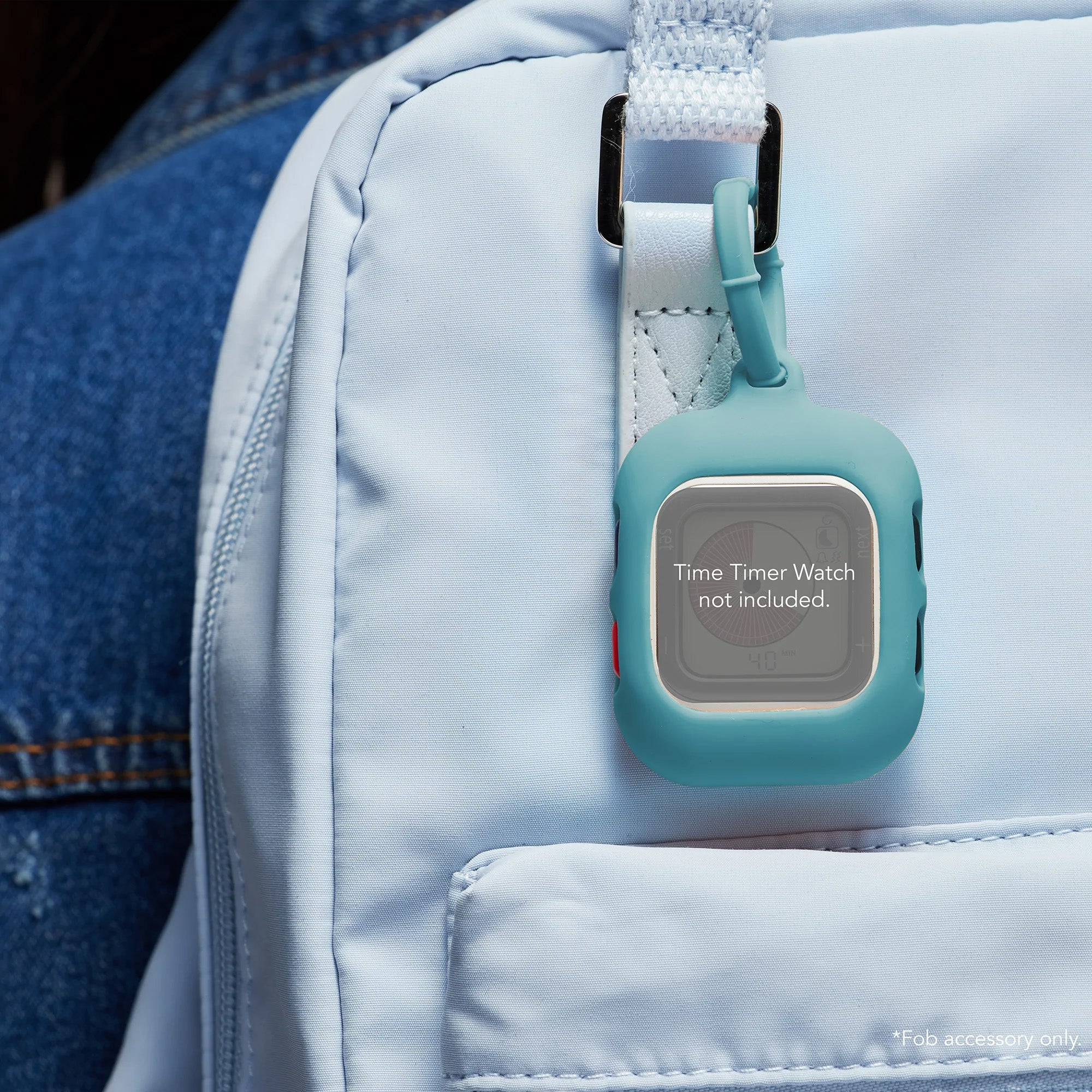 A blue Time Timer Watch Fob Case by Time Timer is clipped to a light blue backpack. The case reads "Time Timer Watch not included." A person in a denim jacket, partially visible, wears the backpack.