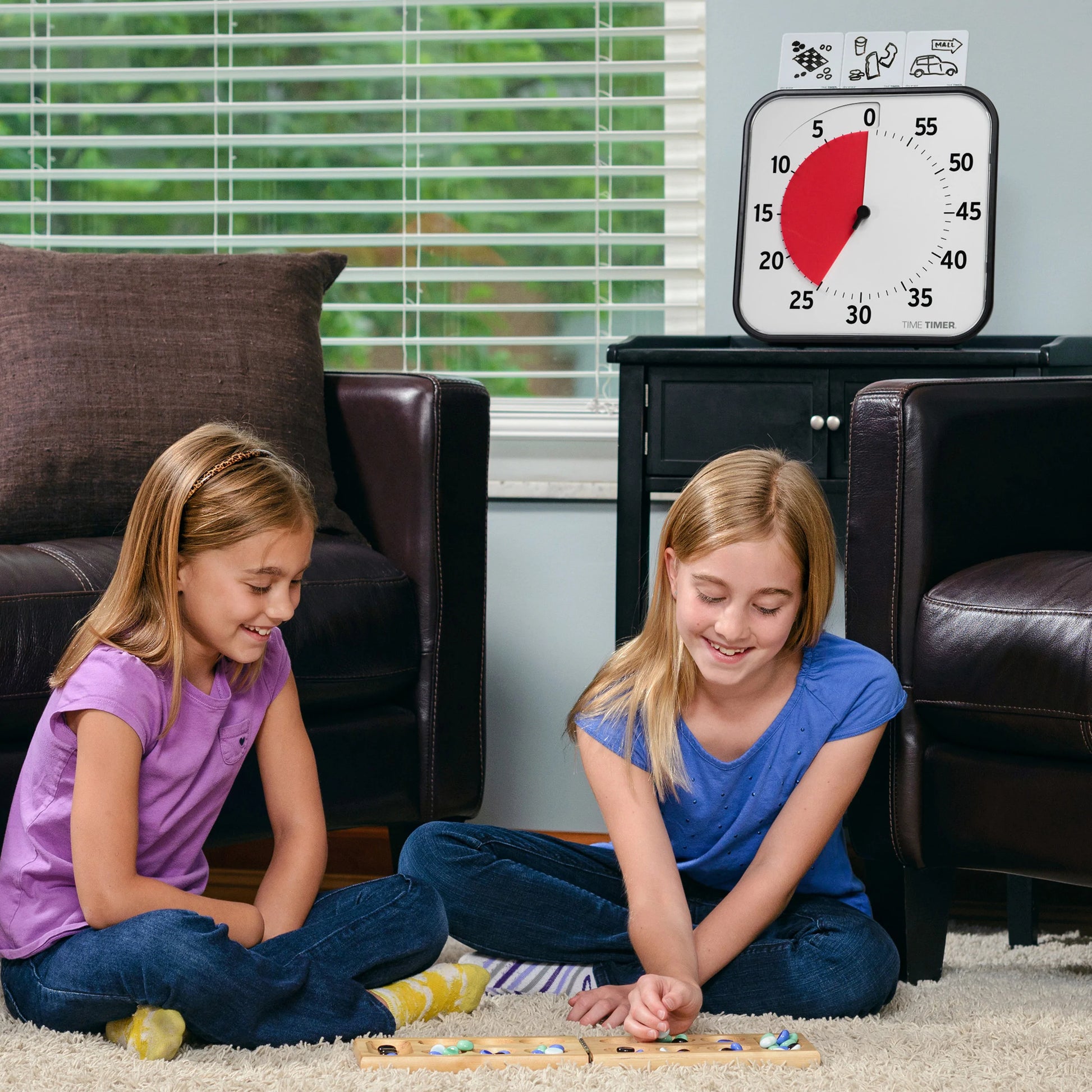 Two young girls sit on the floor playing a board game in the living room. Behind them are two brown chairs, a window, and a Time Timer Original (12 Inch) by Time Timer on the wall showing about 45 minutes left.