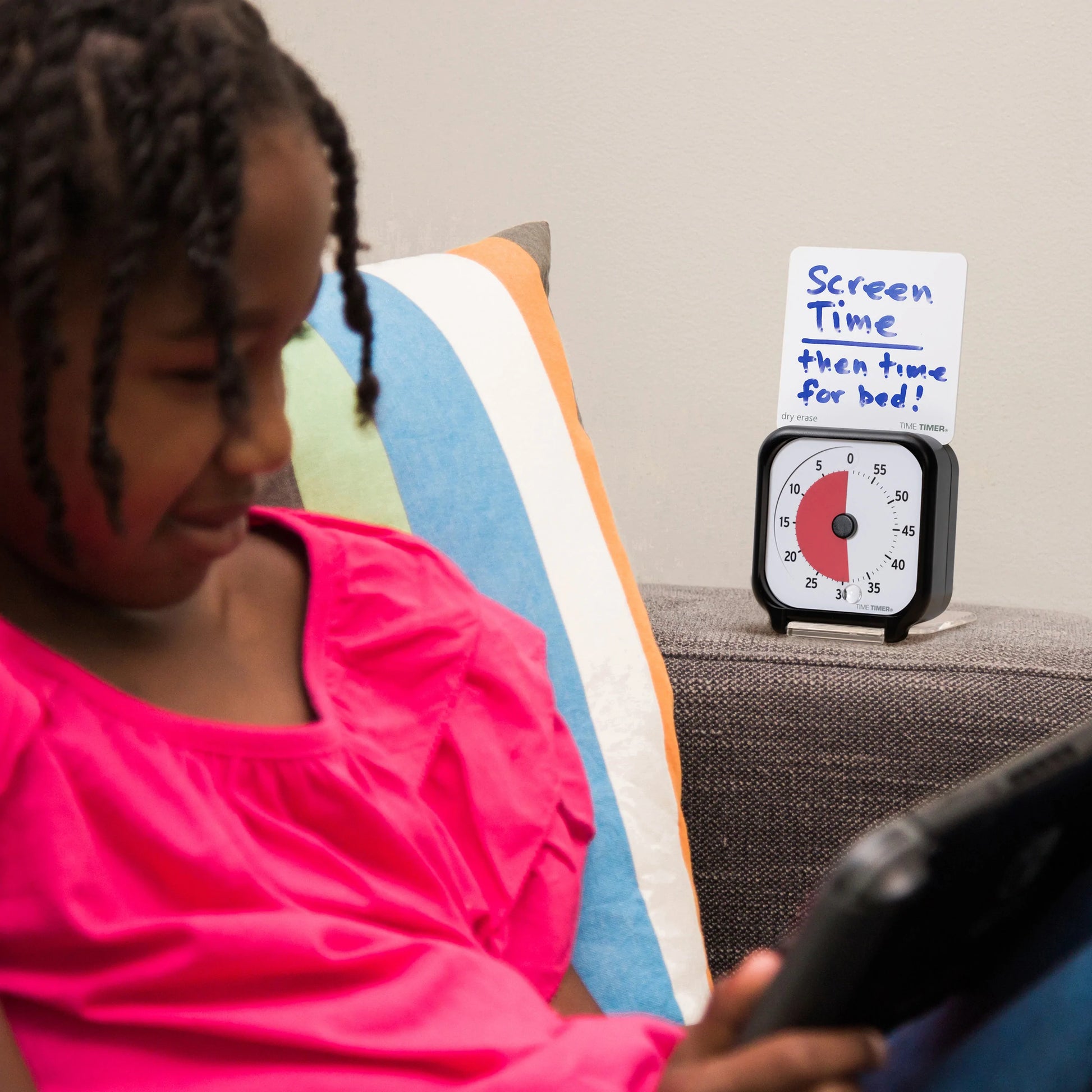 A young girl in a pink shirt uses a tablet on the couch. Beside her is the Time Timer Original (3 Inch) Pocket by Time Timer, showing about 15 minutes left and a note: "Screen time then time for bed!"—ideal for teaching time management.