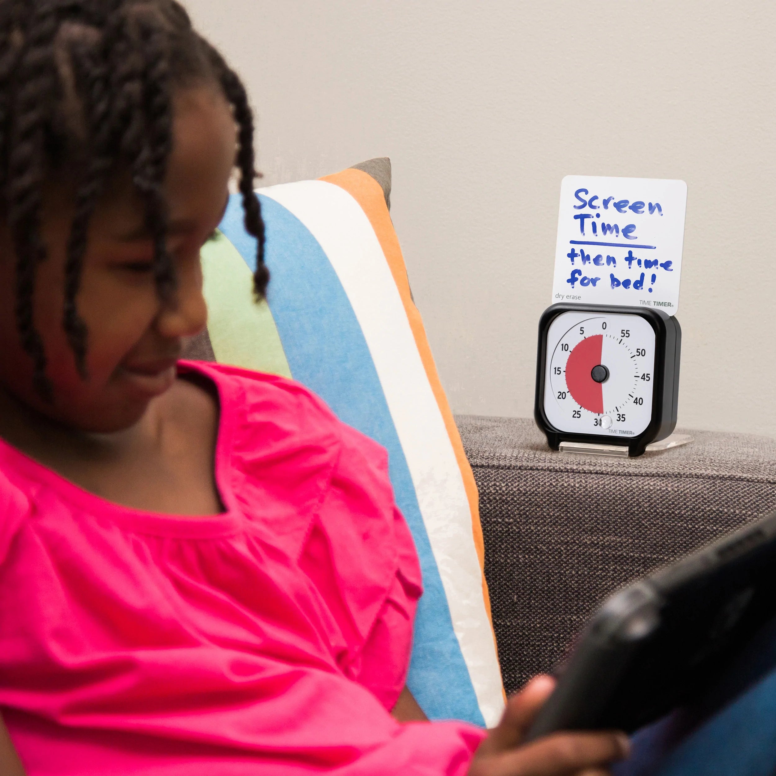 A young girl in a pink shirt uses a tablet on the couch. Beside her is the Time Timer Original (3 Inch) Pocket by Time Timer, showing about 15 minutes left and a note: "Screen time then time for bed!"—ideal for teaching time management.