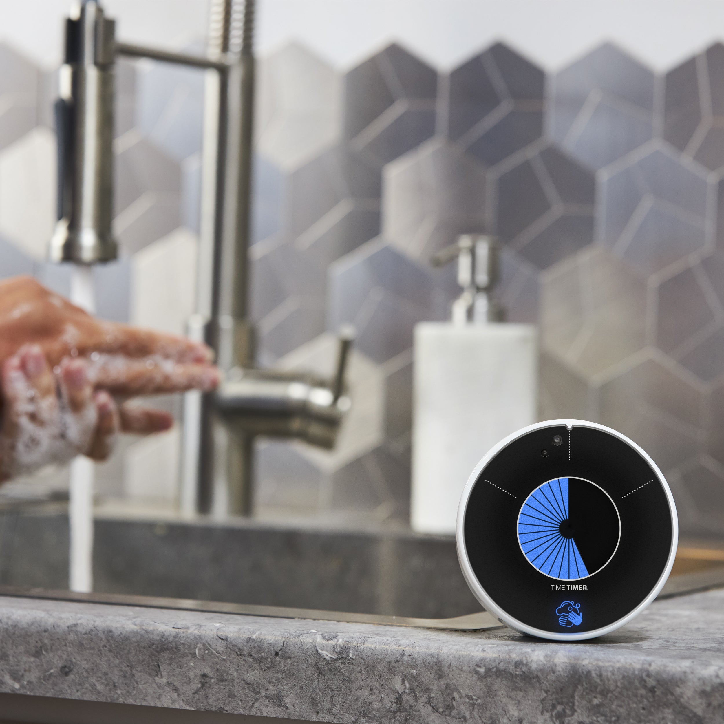 A person washes hands at a kitchen sink, while the Time Timer PRO Applied Touchless 30 Second Handwashing Visual Timer by Time Timer counts down in blue on the countertop. A soap dispenser and metallic backsplash are also visible.